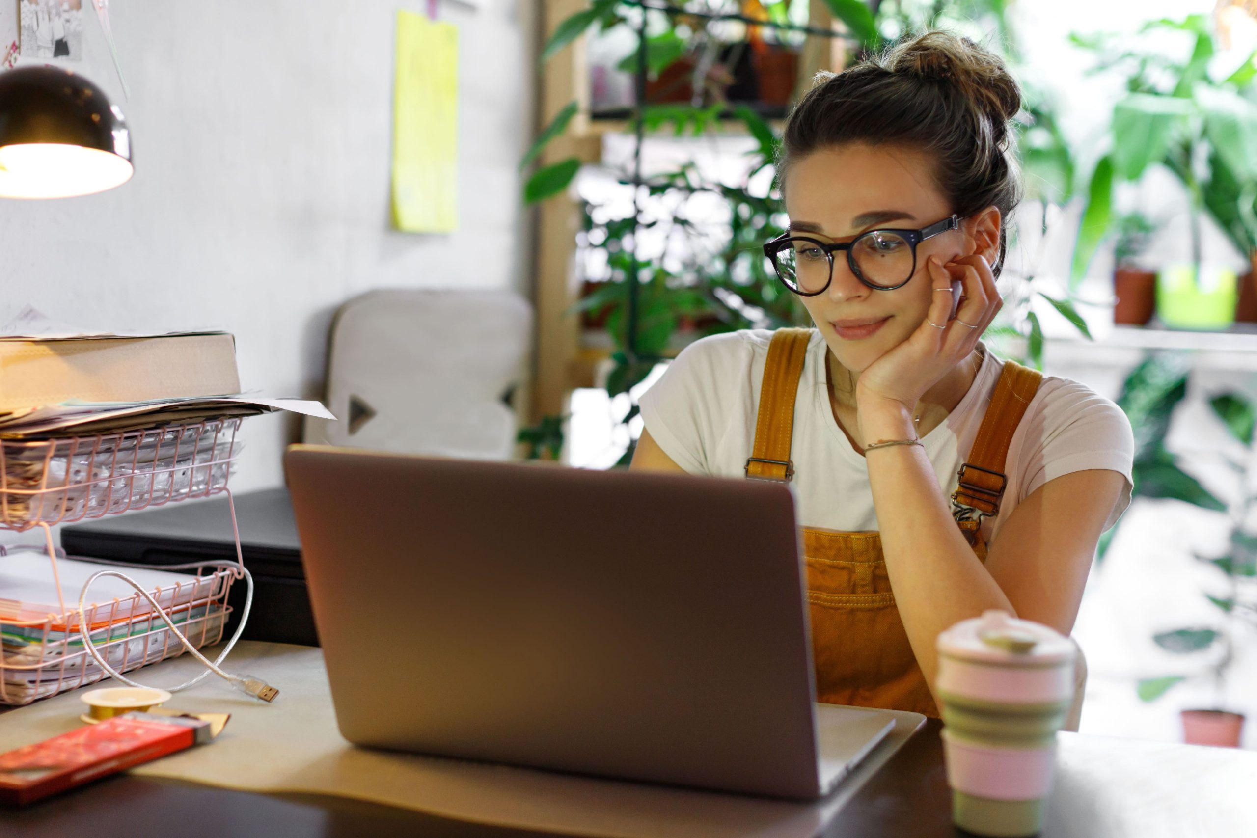 Woman with glasses working on laptop at home, surrounded by plants. Flexible working concept.