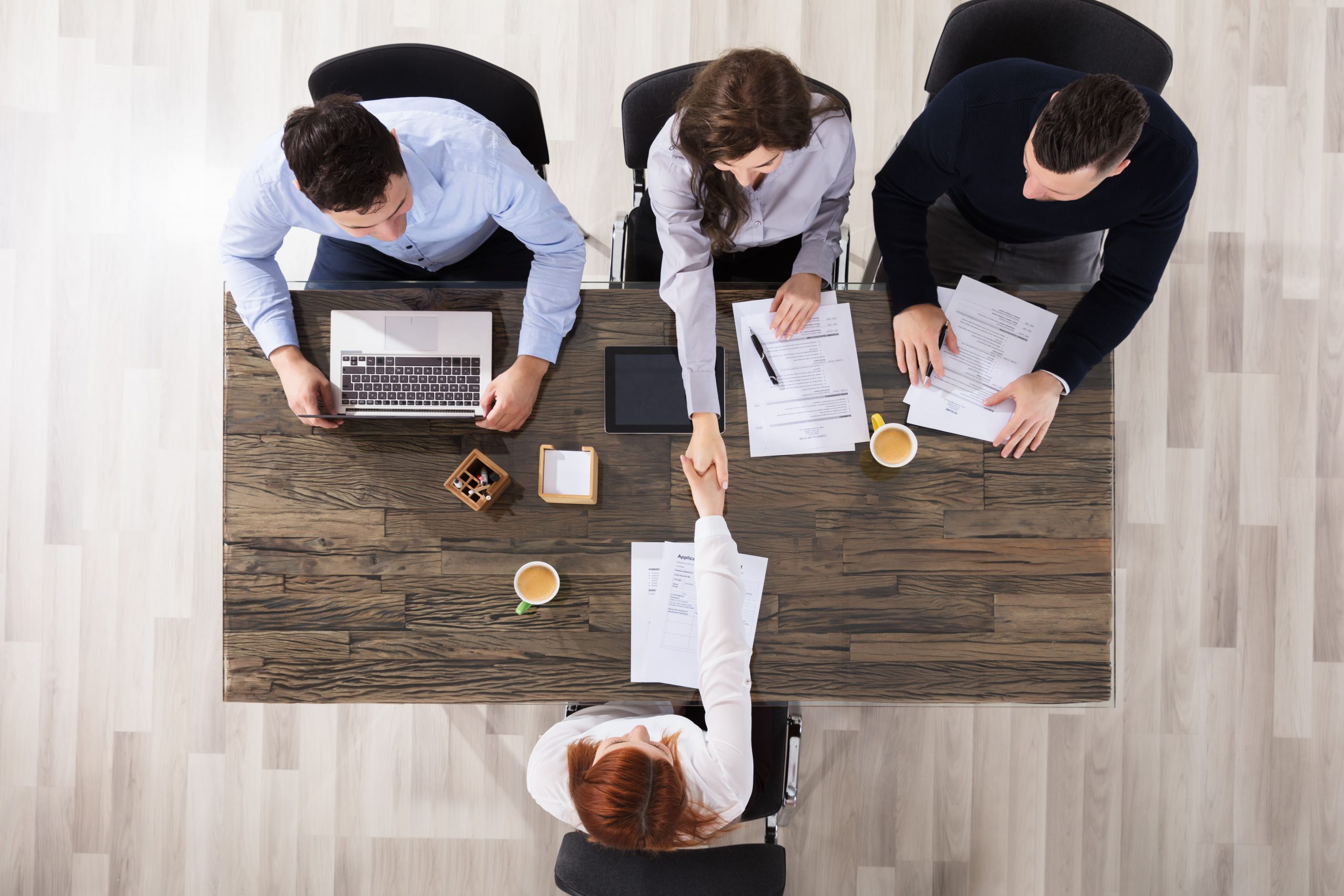 Job interview: Candidate shaking hands with recruiters across a wooden table with documents and a laptop.