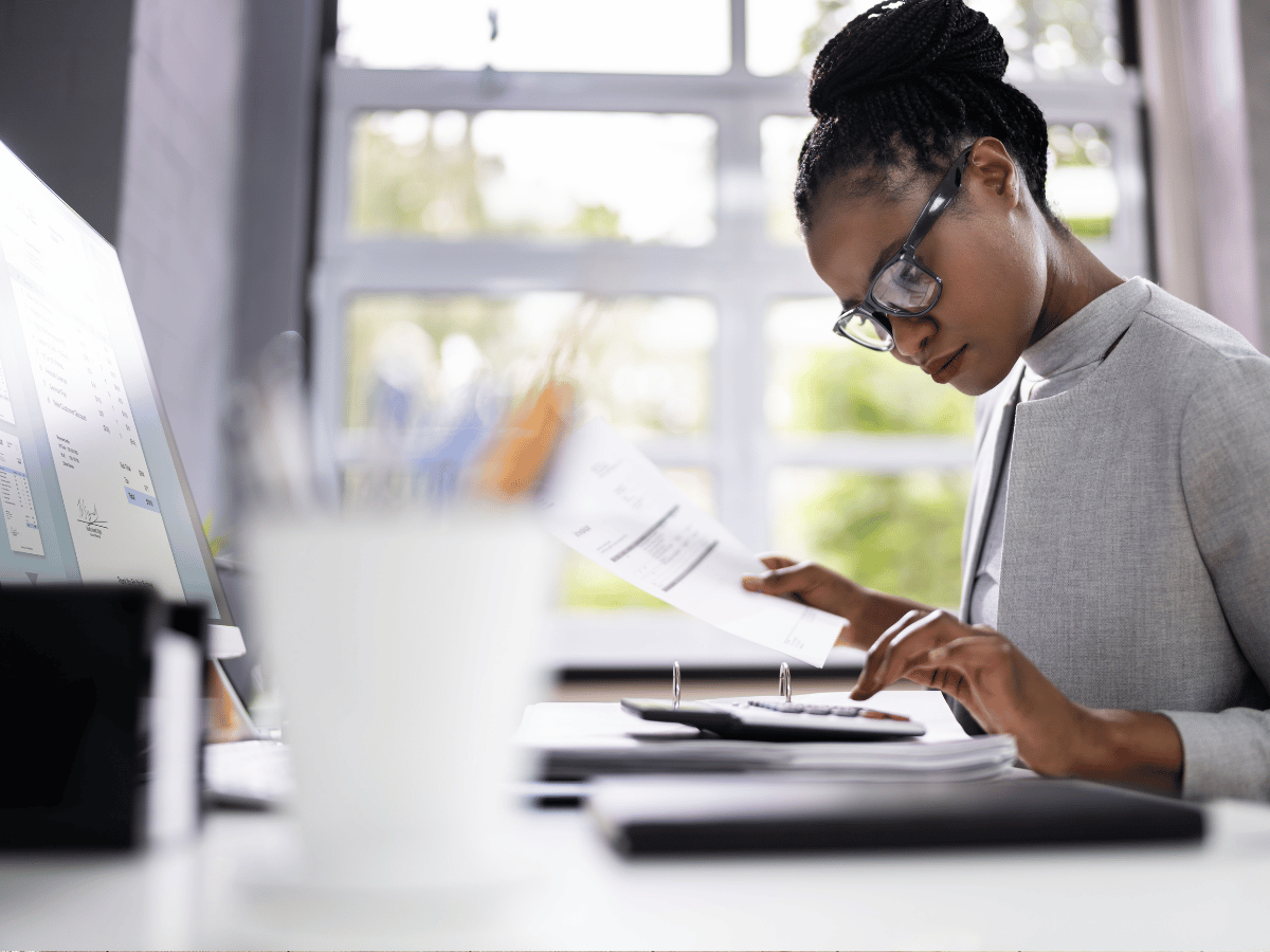 A woman in glasses sits at her desk studying for accountancy exams after recruitment. She holds a sheet of paper and is using a calculator.